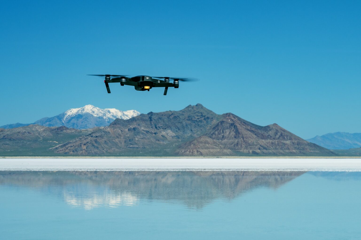 A drone flys over a reflective like with mountains and a clear blue sky in the background