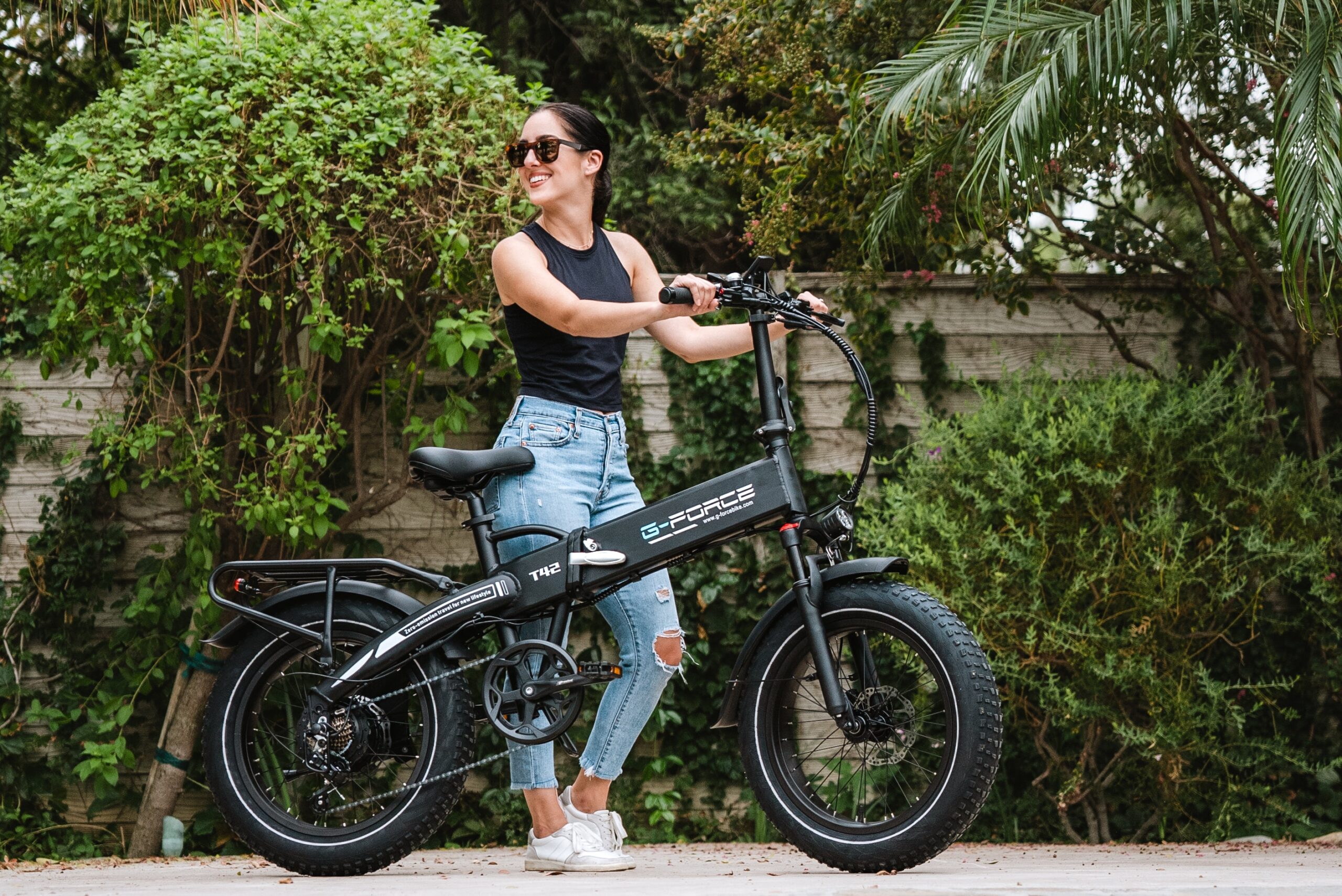 A woman holding an electric bike with bushes, trees and a fence behind her