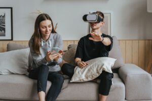 A boy and a girl sat on a sofa with the boy wearing a VR headset and a girl holding an iPhone