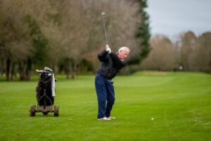 A man on a golf course about to swing stood next to a gold trolley