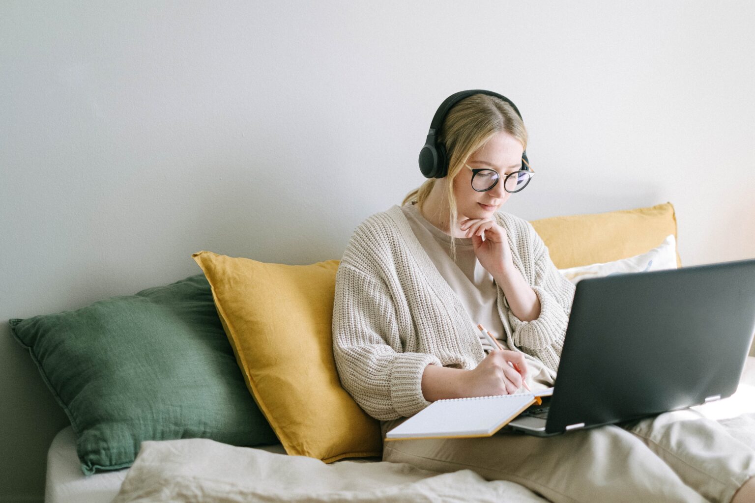 A woman wearing glasses sat on the sofa using her laptop and writing in a notebook with wireless headphones on