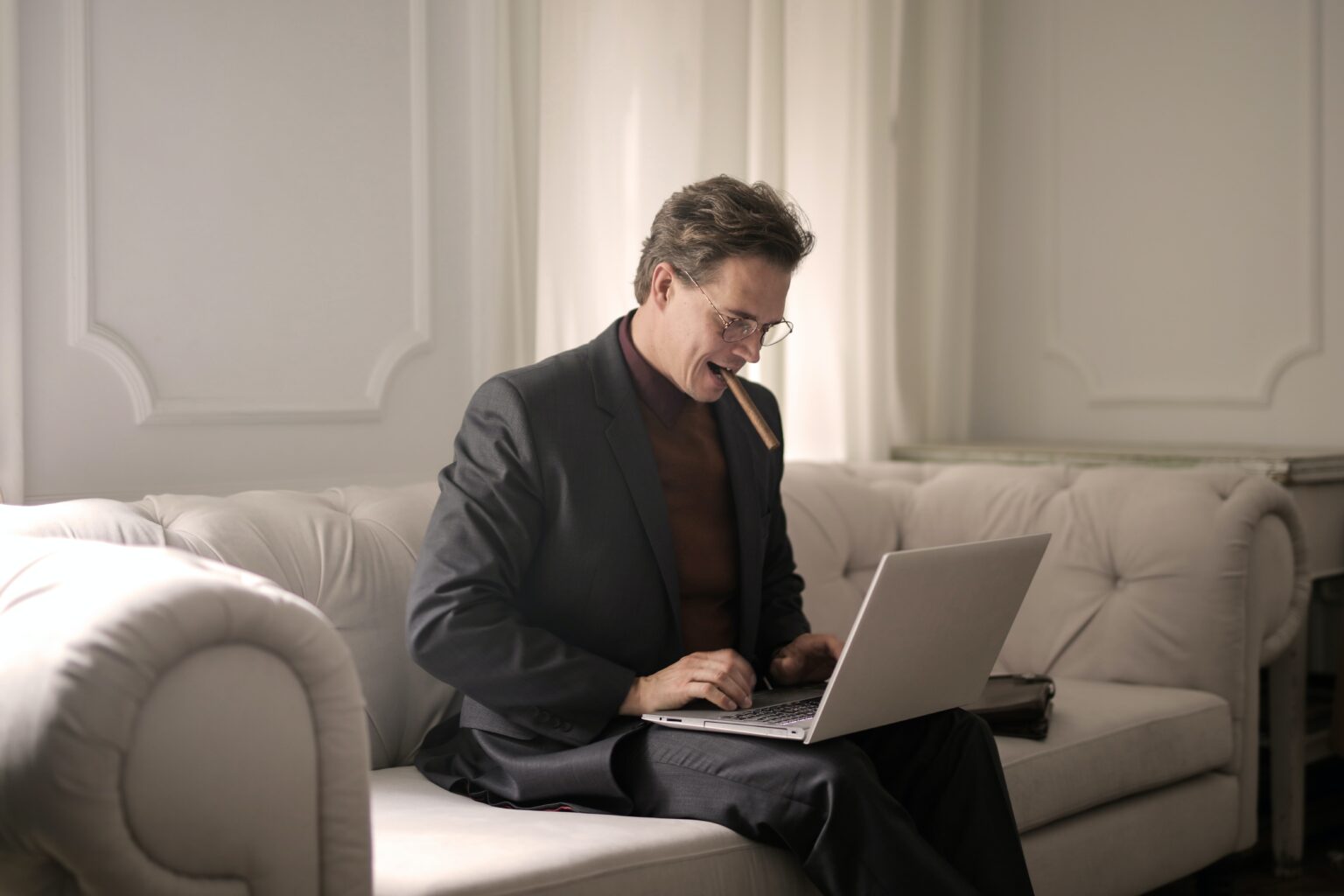 A man in a suit sat on the sofa using a laptop with a cigar in his mouth